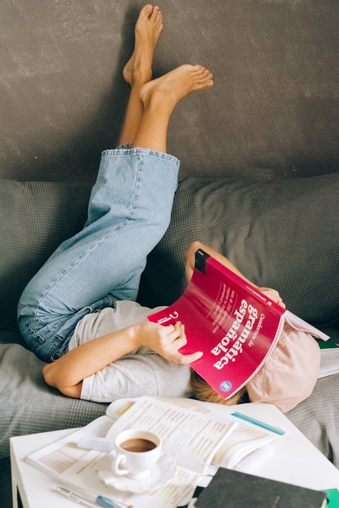 Young woman relaxes upside-down on a couch while studying a Spanish book indoors.